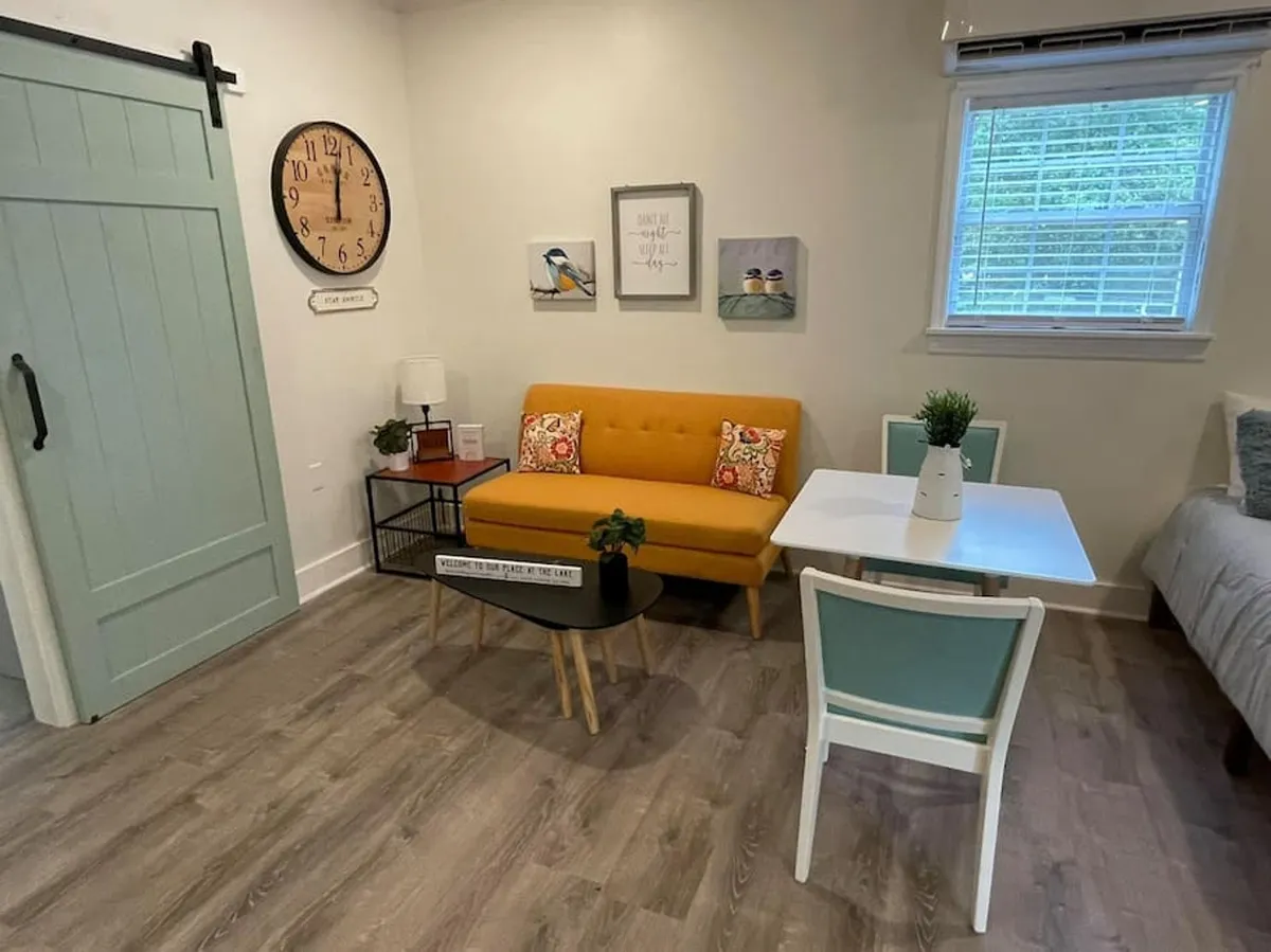 A sunlit studio living area featuring a bright yellow sofa, black coffee table, wall art, and a green accent chair. The large window with blinds allows natural morning light to fill the space, making it feel open and inviting. The arrangement offers multiple places to sit or set up a laptop, highlighting the room's versatility.