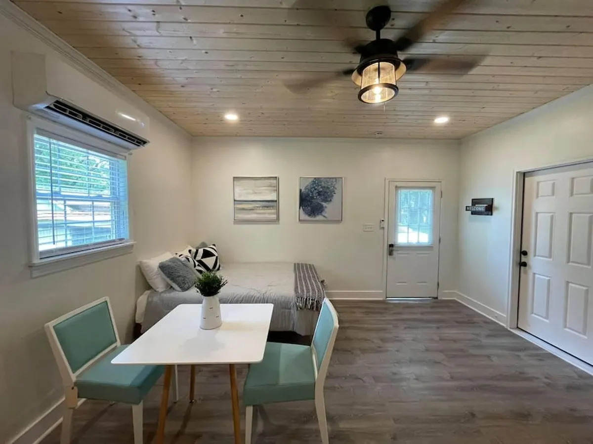 This image features a cozy sleeping area with a bed adorned with decorative pillows, a small dining table nearby, and artwork on the wall. The natural light filtering into the space creates a tranquil and inviting atmosphere, perfect for unwinding after a long day or waking up refreshed.