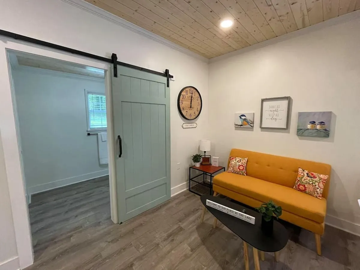 Another view of the same welcoming living space, this time highlighting the yellow sofa from a different angle, complemented by a rustic barn-style sliding door, a decorative wall clock, and curated wall art. The overall mood is warm and homey, with elements that suggest both relaxation and creative inspiration. It feels like a great spot to read, work remotely, or have a casual conversation.