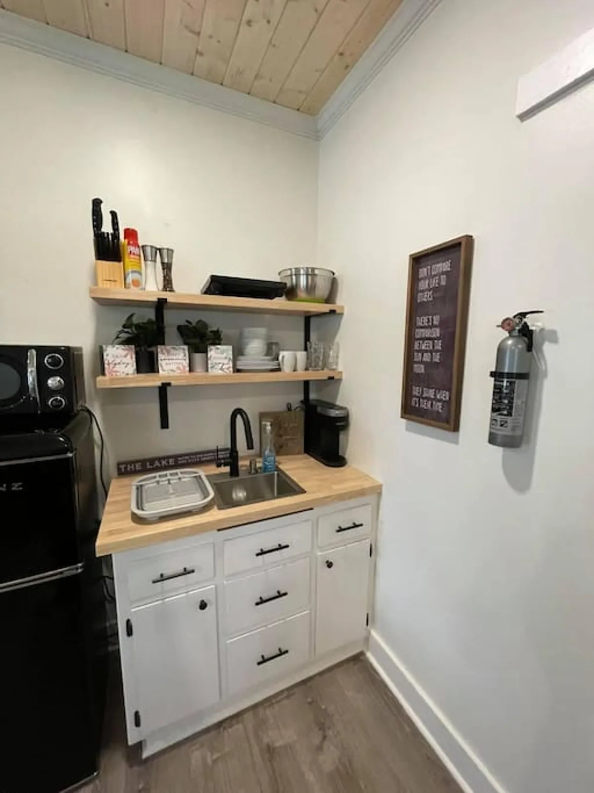 This image features a compact kitchenette with a well-organized sink area, open shelves stocked with essentials, a coffee maker ready for morning brews, and a decorative sign that adds a welcoming touch. The scene is practical and inviting, illustrating meal preparation and a space where guests can socialize or enjoy coffee together.