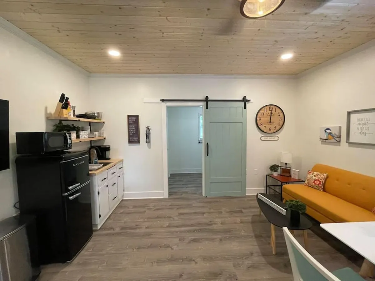 This image features a vibrant orange couch, a striking green sliding door, a black refrigerator, and beautiful wooden ceilings. The combination of the inviting sitting area and architectural details imparts a cozy, welcoming atmosphere. Natural light gently streams in, highlighting the textures and warmth of the wood ceiling.