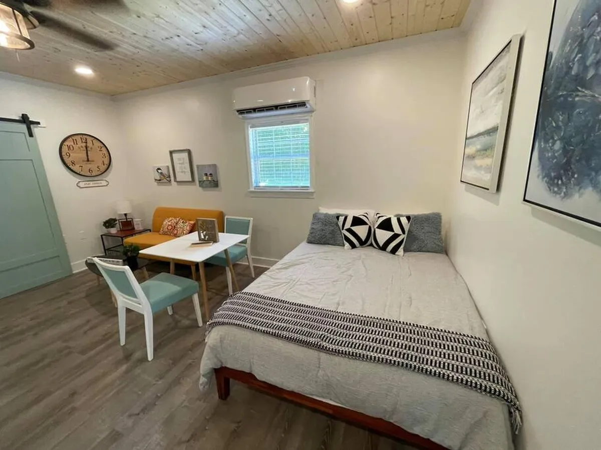 A studio nook with a comfortable bed, a small table and chairs by the window, and tasteful wall art. The space is compact but thoughtfully designed, offering both relaxation and a spot for focused work or dining. The overall mood is cozy and conducive to both concentration and rest.