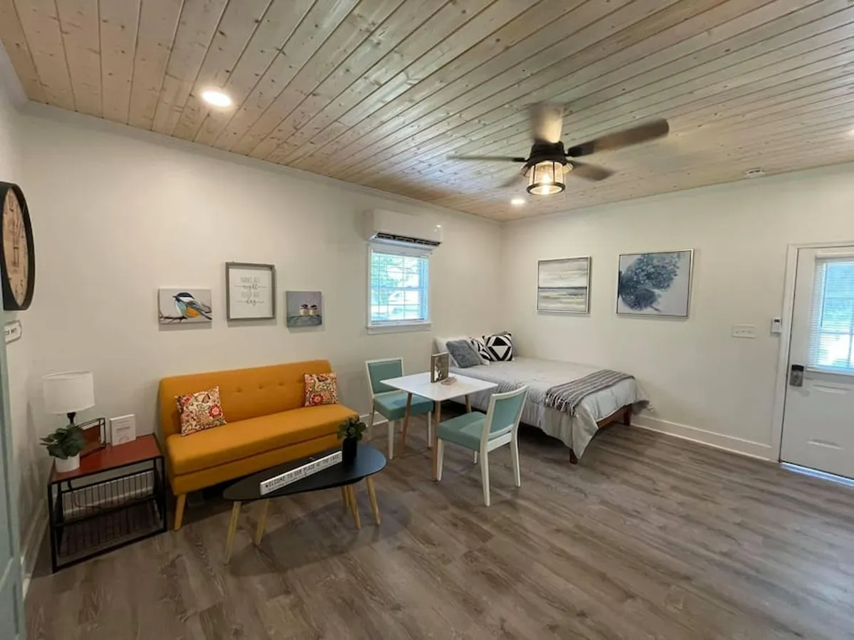 The image shows a bright, inviting studio interior featuring a cheerful yellow sofa, a dining table for two, tasteful artwork adorning the walls, and warm wooden ceilings. Sunlight streams through the windows, creating a cozy ambiance perfect for relaxing, dining, or working remotely.