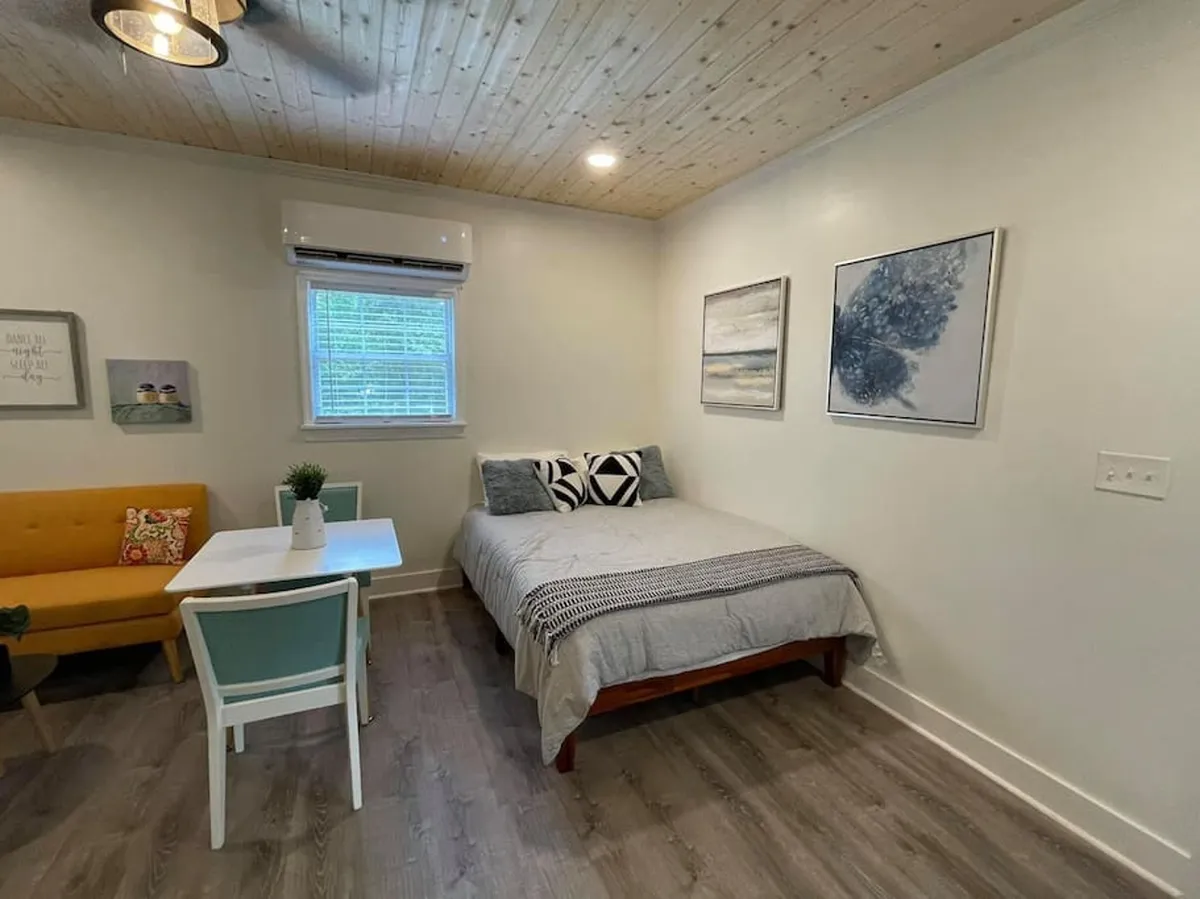 A broader interior view that includes the bed, a sofa, dining table, and prominent artwork. The serene, comfortable mood of the room is evident, with space for both relaxation and socializing. The image showcases the overall atmosphere of peace and comfort.
