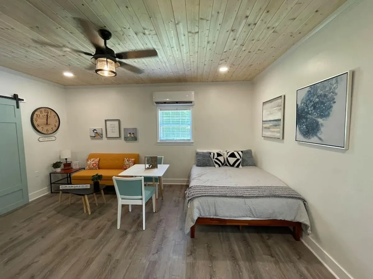 A modern living space with a stylish sofa adorned with vibrant cushions, a well-set dining table nearby, a bed with decorative pillows, and eye-catching wall art. The area feels airy, well-lit, and thoughtfully decorated, blending comfort and style.