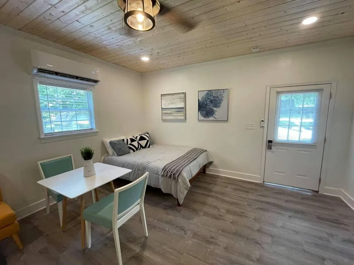 A welcoming interior shot highlighting a bed with decorative pillows, a dining table with chairs, and art-filled walls. The door leading outside hints at privacy and a peaceful retreat within the sleeping area, while the overall setup suggests comfort and relaxation.