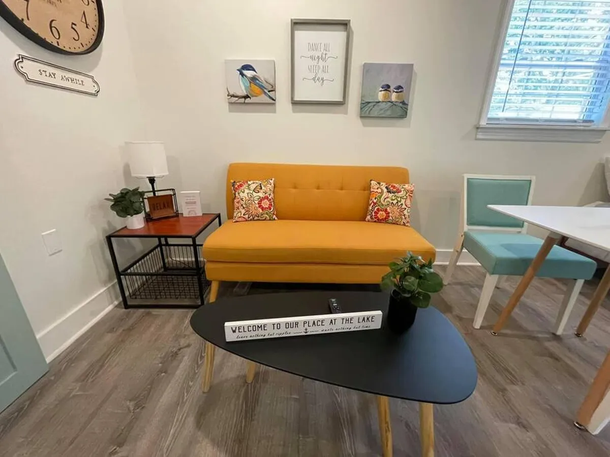 A bright, inviting living area featuring a cheerful yellow sofa adorned with colorful cushions, centered around a black coffee table with a decorative sign. Vibrant wall art adds personality, and the space is bathed in natural light, creating a comfortable environment ideal for relaxing, socializing, or enjoying a cup of coffee. The overall effect is cozy yet modern, perfect for unwinding after a work session or gathering with friends.